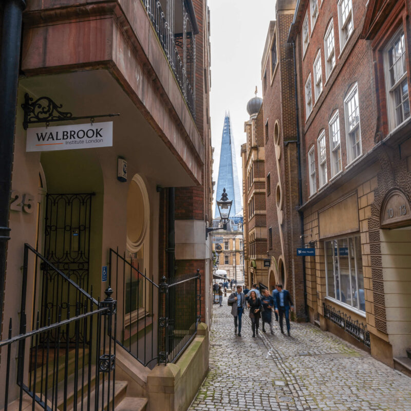 Image of a narrow street in central London. You can see the Shard through the gap at the end of the street. A hanging sign on a building on the left hand side reads "Walbrook Institute London"