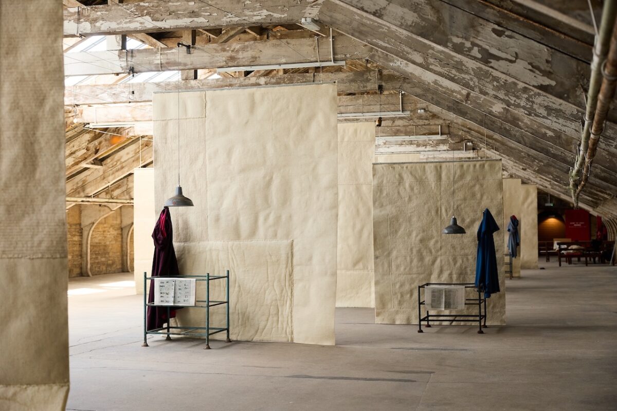 An art installation hangs in a large industrial space with exposed wooden rafters. Large beige textile pieces are suspended from the ceiling to the floor which fill the room.