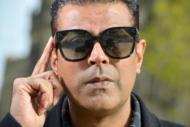 Zanco CEO Shazad Talib models the black smart glasses outside on a sunny day. He holds his right hand up to the arm of the glasses as if to adjust a setting.