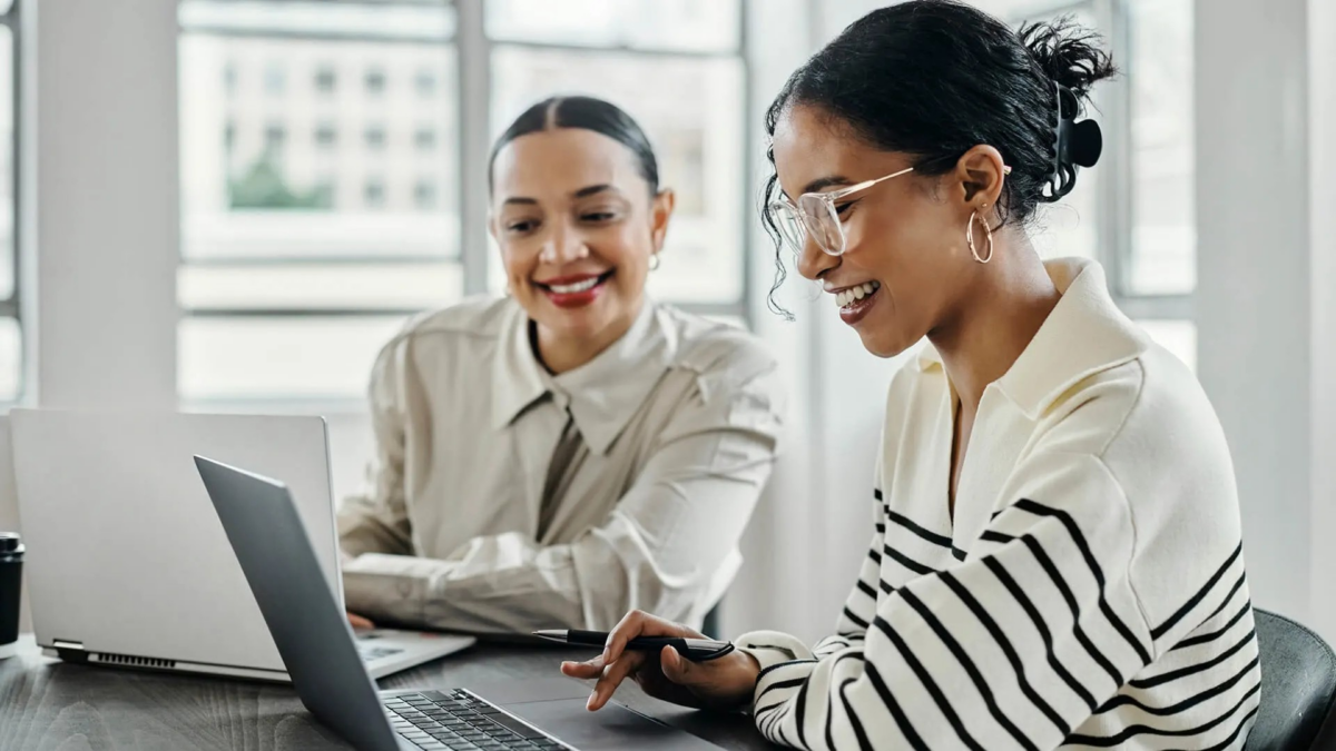 Two young women are looking at laptops in a study or work environment with a window behind them.